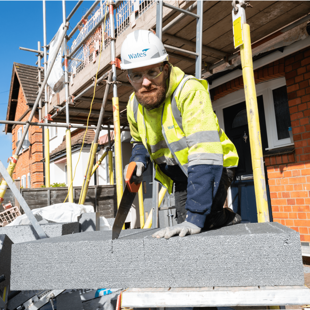 An individual who is wearing a yellow hi-vis jacket and black work trousers is using a saw to cut through a grey insulation slab. In the background, there is a brick house that is undergoing construction.