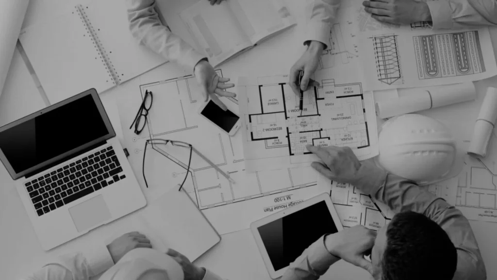 A group of professionals collaborating on a retrofit design project. The image shows architectural blueprints, a laptop, a tablet, and a hard hat on the table, with hands pointing at retrofit plans. This represents the role of a Retrofit Coordinator in planning and executing energy-efficient building upgrades.