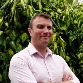 An individual called Adrian Marshall is wearing a pink shirt whilst posing in front of a green tree.