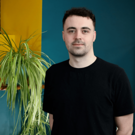 An individual called Anthony Mcgill has short black hair and beard whilst wearing a black t-shirt and is posing in front of a green and yellow wall with a plant next to them.