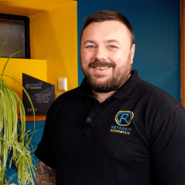 Callum Harrison a member of Retrofit Academy is wearing a black Retrofit branded polo t-shirt and is standing in front of a blue wall.