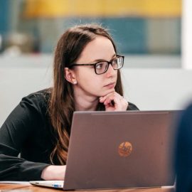 An individual called Charlotte Salt is sat at a desk wearing a black shirt with glasses and has a laptop in front of them.