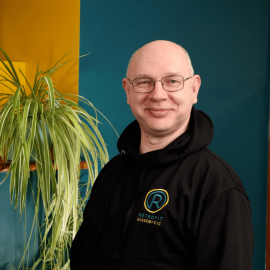 An individual called Clive Rowland is wearing a branded Retrofit black hoodie whilst posing in front of a blue wall next to a green plant.