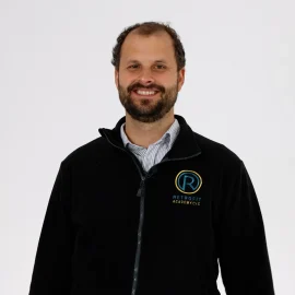 A photo of an individual called Dom Ives who has short brown hair and a beard is wearing a black Retrofit Academy jacket whilst smiling at the camera.