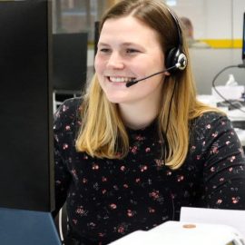 Ellie Thomas is sat at a desk wearing a pink and black shirt with a headset on whilst looking at a computer screen.