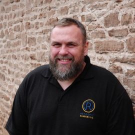 Gary Rivett is wearing a branded Retrofit black polo t-shirt whilst smiling at the camera and is standing in front of a brick wall.