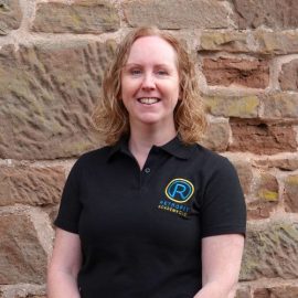 Jenny Pierpoint is wearing a black Retrofit polo t-shirt and is standing in front of a wall.