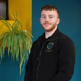 A photo of Josh, a member of the Retrofit Academy team, is wearing a branded black Retrofit Academy zip up jacket, has short brown hair with brown facial hair and is standing in front of blue and yellow walls with a green plant next to him.