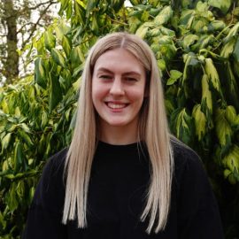 An individual called Lucy Postle has long blond hair and is wearing a black jumper whilst posing in front of a green tree.