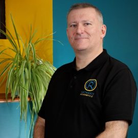 An individual with short hair is wearing a black Retrofit branded polo shirt whilst posing in front of a blue wall next to a green plant.