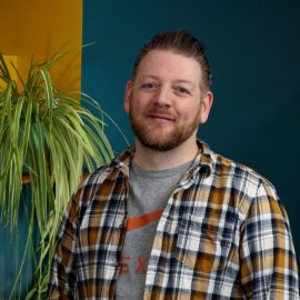 An individual called Matt Elliot is wearing a checked shirt whilst posing in front of a blue wall next to a green plant.