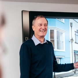 A photo of an individual who has short grey hair, is wearing a dark blue jumper and a light blue shirt, is smiling at a camera.