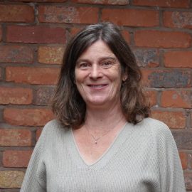 An individual called Susan Gregory has long brown hair whilst wearing a grey jumper and is posing in front of a brick wall.