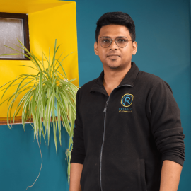 An individual with dark hair and glasses is wearing a black retrofit jacket and is standing in front of a blue wall.