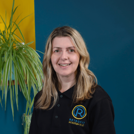 An individual whop has long brown hair is wearing a black Retrofit branded polo t-shirt whilst posing in front of a blue wall and next to a green plant.