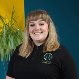 An individual who has long blond hair is wearing a black Retrofit branded polo t-shirt whilst posing in front of a blue wall and next to a green plant.