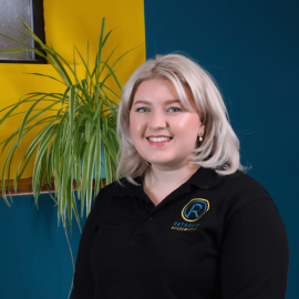 An individual who has long blond hair is wearing a black Retrofit branded polo t-shirt whilst posing in front of a blue wall and next to a green plant.