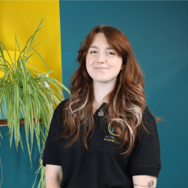 An individual with long brown hair is wearing a Retrofit branded black polo t-shirt whilst posing next to a green plant, in front of a blue wall.