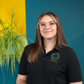 An individual with long brown hair and glasses is wearing a Retrofit branded black polo t-shirt whilst posing next to a green plant, in front of a blue wall.