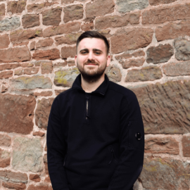 An individual with brown hair and a brown beard is wearing a black jumper and is standing in front of a brick wall.