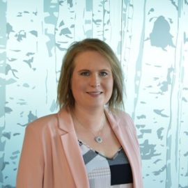 An individual who has short shoulder length brown hair is wearing a pink blazer with a black and white patterned top and is standing in front of a wall with a fake tree pattern.