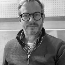 An individual who has short grey hair and beard is wearing a jumper whilst smiling at the camera.