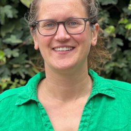 An individual called Lizzy Westmacott is standing in front of a green bush whilst wearing brown glasses and a bright green shirt.