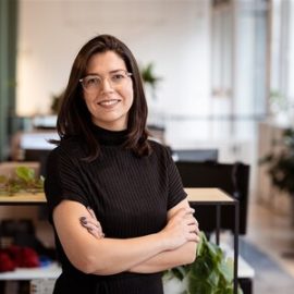 A photo of an individual who has long brown hair, clear glasses and a long black shirt, whilst posing in an office environment.