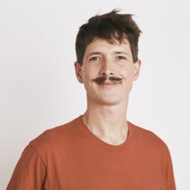 An individual who has short brown hair and a moustache whilst wearing a light orange t-shirt is smiling at the camera.